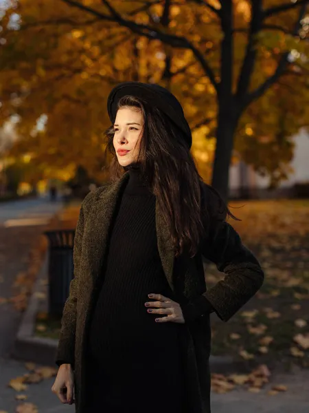 年轻美丽的孕妇 头戴黑发 身穿黑色紧身衣 头戴一顶帽子 躺在公园里秋天的草地上 图库图片