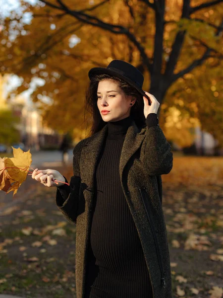 年轻美丽的孕妇 头戴黑发 身穿黑色紧身衣 头戴一顶帽子 躺在公园里秋天的草地上 免版税图库照片