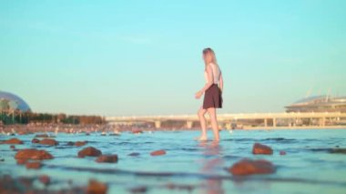 Kahverengi etekli, beyaz tişörtlü, sıska, güzel, Avrupalı bir kız akşam vakti taşlarla kıyıda yürüyor ve Saint-Petersburg 'daki Gazprom Stadyumu' nda açık mavi arka planda Gazprom Stadı var.