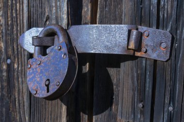 Eski, paslı bir ahır kilidi ahşap bir kapının desteğinde asılı. Yan görüş. Yakın plan. Zor gün ışığı.