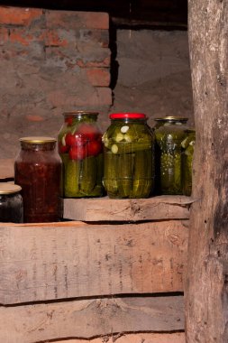 Konserve sebzeli cam kavanozlar (salatalık ve domatesler) kırsal bir mahzendeki ahşap bir rafta bir sırada dururlar. Tozla kaplı teneke kutular. Tahtalar ön planda. Tuğla duvar Yakın plan..