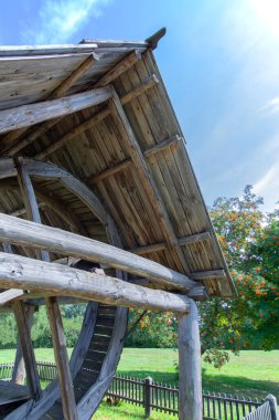 Çiçekli dağ küllerinin arka planında tekerlek olan eski bir ahşap kuyunun manzarası. Gökyüzü açık. Yaz. Gün ışığı.