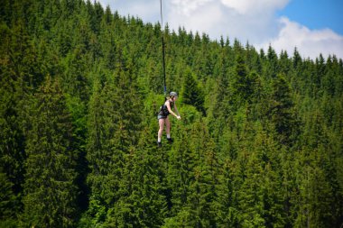 Kız bungee jumping binişinden atladı, yüksek irtifada sıkı bir ipe tutunup gülümsedi. Bir dağ yamacının bulanık arka planında