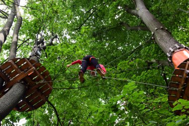 Emniyet kemeri takmış genç bir çocuk halat parkındaki asma köprüden geçiyor.