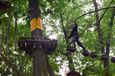 Genç bir çocuk bir ağaçtan diğerine merdiven asarak lunaparkta tırmanır.