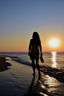 Genç bir kadın deniz kenarında çıplak ayakla yürüyor, elinde ayakkabıları var. Sadece siluet görünüyor. Arka plan