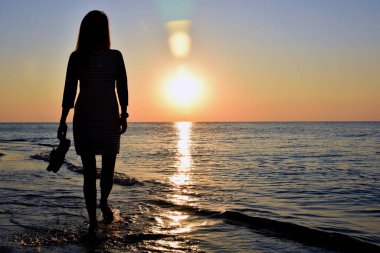 Genç bir kadın deniz kenarında çıplak ayakla yürüyor, elinde ayakkabıları var. Sadece siluet görünüyor.
