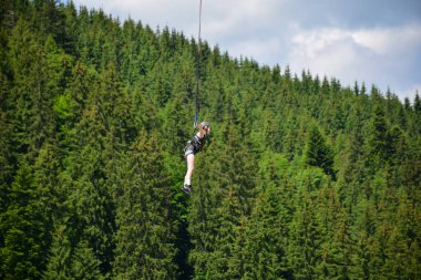 Kız bungee jumping gezisinden atladı, yüksek irtifada sıkı bir ipe tutundu. Bir dağ yamacının bulanık arka planında