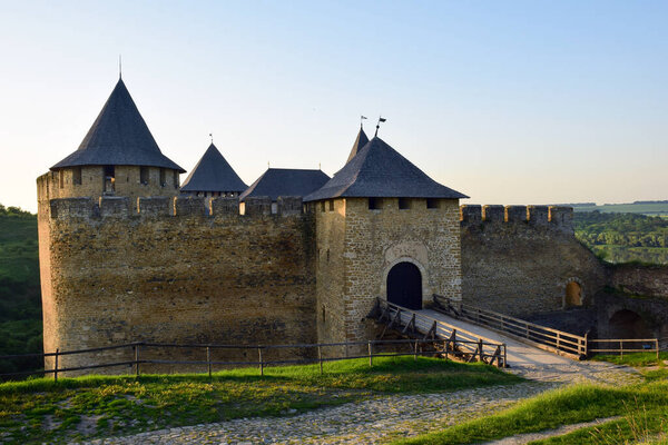Towers of a medieval stone fortress with battlements, an arched entrance and a wooden bridge, and a cobbled road to it