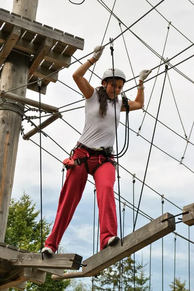 İp eğlence parkındaki genç bir kadın koşum takımı içinde asma ahşap bir köprüden geçiyor.