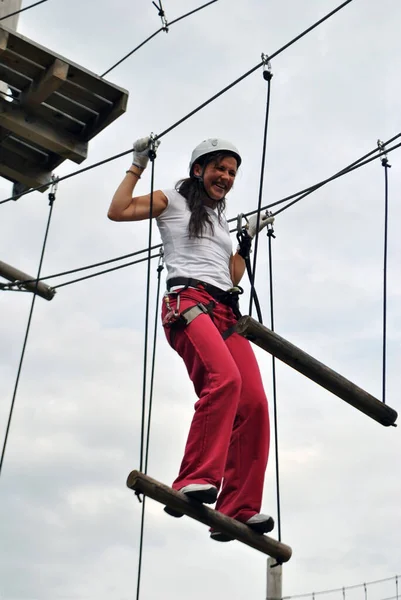 Eğlence parkında genç bir kadın emniyet kemeri ve kahkahalarla ip köprüsü boyunca yürüyor.
