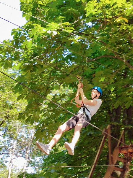 Bir genç halatla bir teleferiğe iniyor. Bir halat parkında, ağaçların arkasına doğru.
