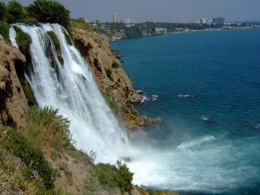 Büyük bir yükseklikten denize düşen suyla birlikte şelale. Arka planda şehri ve kayaları görebilirsiniz.