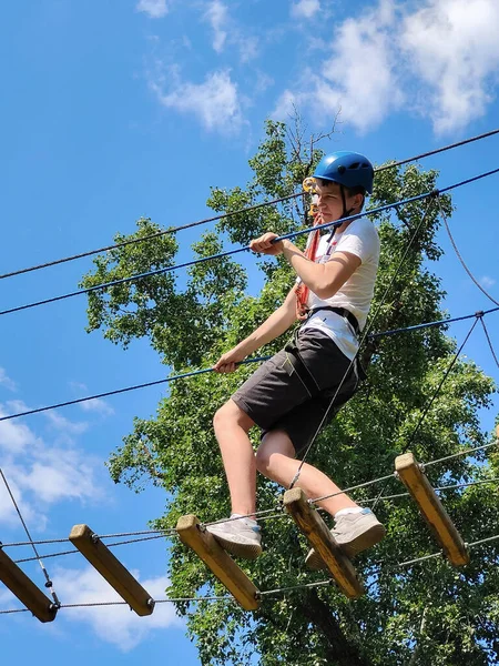 A teenager boy walks along a rope bridge between trees in an amusement park in safety gear and a helmet. Photo from the bottom