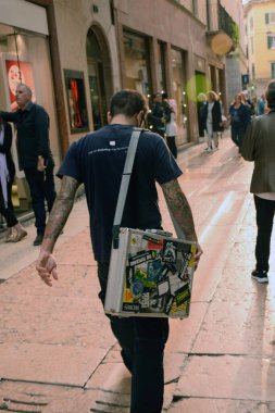 Omzunda çıkartmalarla dolu bir bavulu olan bir sanat adamı sokakta insanların arasında yürüyor. Arka plan