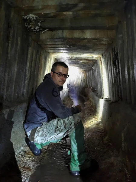 A  man, a digger, crouched in the underground tunnel of the sewage collector in perspective. A stream flows under it