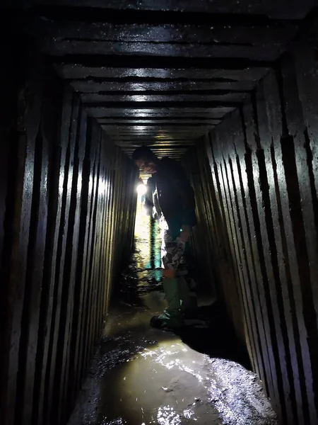 A young man, a digger, crouched in the underground tunnel of the sewage collector in perspective. A stream flows under it