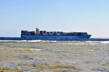 A huge container ship is carrying many commercial containers against a clear blue sky. Side view from the shore
