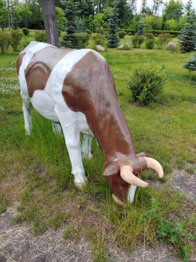 A statue of the figure of a spotted cow in the park as a life-size decoration on a background of grass and trees