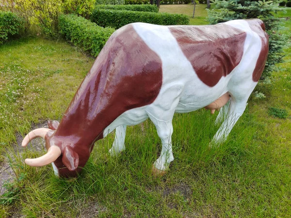 A statue of the figure of a spotted cow in the park as a life-size decoration on a background of grass and trees