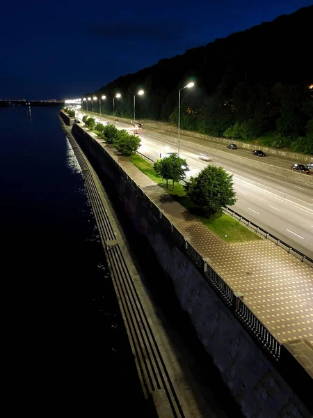 The night embankment near the river in the perspective of illumination by street lamps. There are few cars on the road