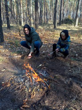 Two teenagers of different ages are sitting by a bonfire in a cold forest and grilling sausages on skewers.