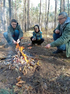 A father and two sons sit by a fire in the forest and grill sausages on skewers
