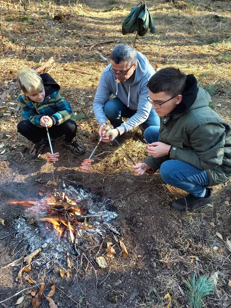 A father and two sons sit by a fire in the forest and grill sausages on skewers