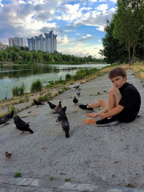 Genç bir çocuk parktaki nehir kıyısında elleriyle güvercinleri besler. Gelecekte yüksek binalar