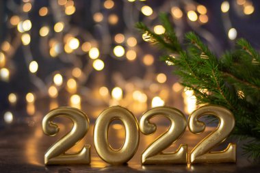 Mutlu yıllar 2022. Altın sayılar, arka planda parlak bir bokeh ve bir ladin filizi bulunan modaya uygun mavi bir zemin üzerinde dururlar..