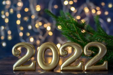 Mutlu yıllar 2022. Altın sayılar, arka planda parlak bir bokeh ve bir ladin filizi bulunan modaya uygun mavi bir zemin üzerinde dururlar..