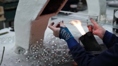 The hands of a glassblower woman holding a glass rod over the flame of a burner against the background of the workplace and tools. Manufacture of Christmas toys made of glass. Craft concept.