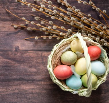 Ahşap bir masanın arka planında, söğüt dalları ve çiçeklerin yanında renkli Paskalya yumurtaları.