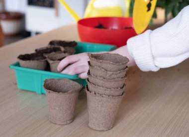 Eve tohum eken genç bir kadının yan görüntüsü. Bir bahçıvan, tahtadan bir masada, yerdeki turba saksılarına tohum eker.