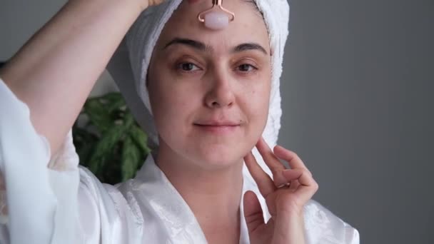 Una foto de cerca de una mujer joven que parece relajada y sonriente haciendo un masaje con un rodillo facial hecho de cuarzo rosa natural. — Vídeos de Stock