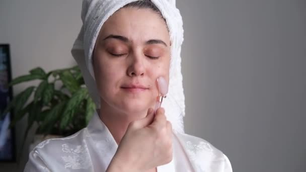 Et nærbillede af en ung kvinde, der ser afslappet og smilende ud med en massage med en ansigtsrulle lavet af naturlig rosenkvarts. – Stock-video