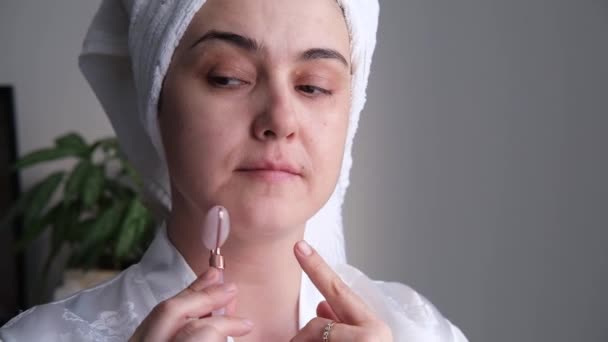 Een close-up foto van een jonge vrouw die er ontspannen en lachend uitziet en een massage doet met een gezichtsrol van natuurlijk rozenkwarts. — Stockvideo