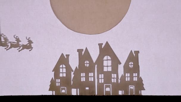 Bábszínház Árnyék Színház. Télapó egy szánon rénszarvas legyekkel a nagy hold előtt a házak felett. A házak ablakai ragyognak. — Stock videók