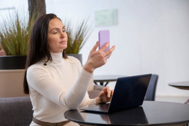 Güzel, orta yaşlı bir kadın açık uzay ofisinde dizüstü bilgisayarının başında oturmuş akıllı telefon yazıyor.