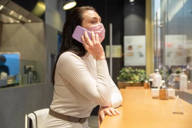 Maskeli kadın kafede telefonla konuşuyor.