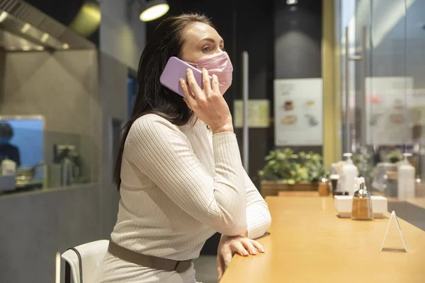 Mujer en máscara hablando por teléfono sentado en la cafetería — Foto de Stock