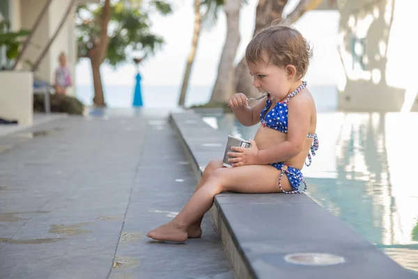 Mała dziewczynka jedząc jogurt lodowy z łyżką siedzi przy odkrytym basenie — Zdjęcie stockowe