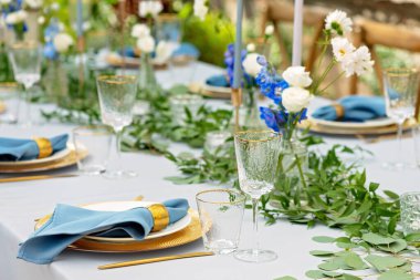 Mavi peçeteli ziyafet masası takımı, altın çatal bıçak takımı, kristal, taze çiçekler ve mumlar. Düğün süslemeleri. Yumuşak seçici odak.