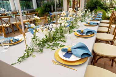 Mavi peçeteli ziyafet masası takımı, altın çatal bıçak takımı, kristal, taze çiçekler ve mumlar. Düğün süslemeleri. Yumuşak seçici odak.