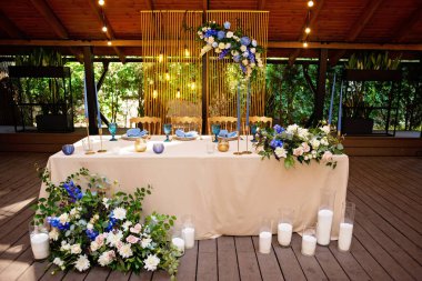 Düğün başkanlığı, çiçekler, yeşillik, mavi ve altın rengi olan yeni evliler için ziyafet masası. Düğün masasında yemyeşil bir çiçek aranjmanı. Parlayan ampuller. Yumuşak seçici odak.