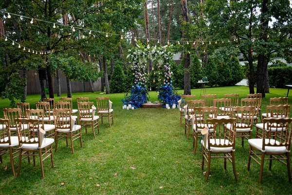 緑の芝生の上で屋外結婚式 白と青の新鮮な花の結婚式のためのアーチとお客様のための金の椅子 柔らかい選択的フォーカス — ストック写真