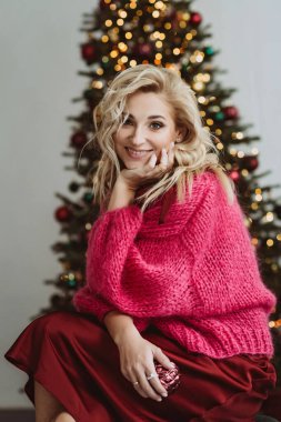 Hoş bir bordo süveter ve ipek bir elbise giyen güzel sarışın kadın, yeni yılı bir Noel ağacının arka planında kutluyor..