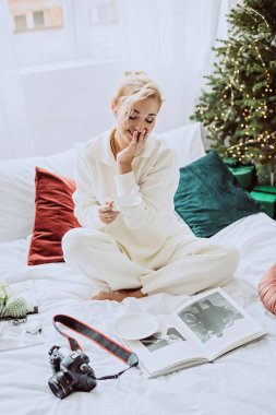 Modern bir yeni yılda, beyaz bir yatak odasında, elinde bir fincan kahveyle bir Noel ağacının arka planında duran genç ve güzel bir kadın. Yumuşak seçici odak.