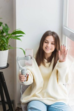 Mutlu genç kız, gülümseyen yüz, el sallayan el, akıllı telefon kamerasıyla konuşmak, video bloğu kaydetmek, sosyal medya yayınlamak, evde video görüşmesi yapmak. Yumuşak seçici odak.