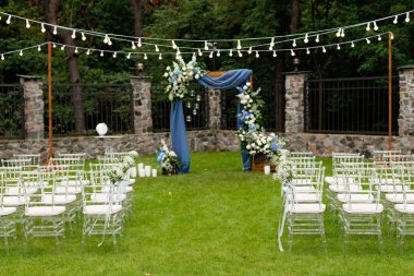 A place for an outdoor wedding ceremony, with a seating area for guests, decorated with chairs. Rustic wooden arch decorated with blue fabric, flower arrangement and candles. Soft selective focus.